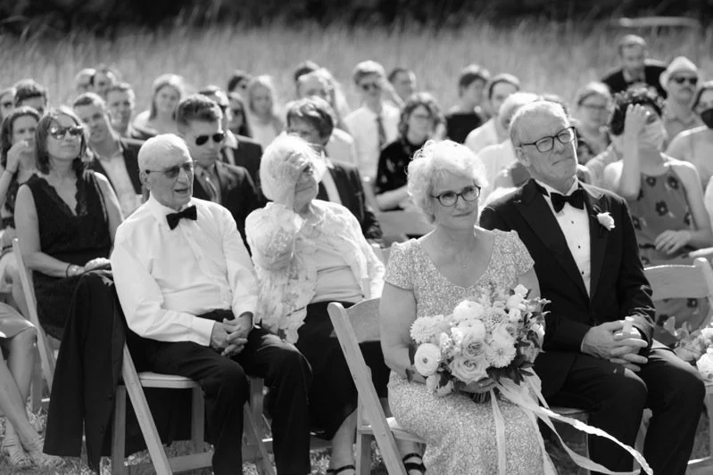 Black-and-white photo of an outdoor wedding ceremony with senior guests sitting in the front row, including a woman holding a bouquet of flowers.
