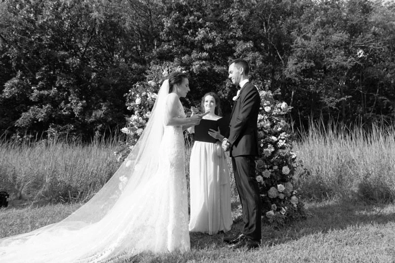 A wedding ceremony outdoors with a bride in a long gown and veil, a groom in a suit, and a officiant reading vows or a speech, with floral decorations and trees in the background.