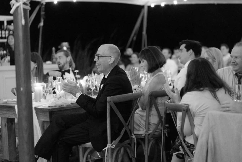 People attending a formal dinner event, seated at tables decorated with candles and glassware, under a tent at night. Some are clapping and smiling.