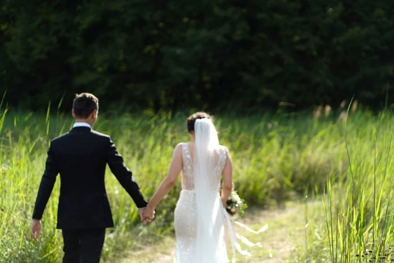 Couple in wedding attire holding hands and walking through a grassy outdoor area.