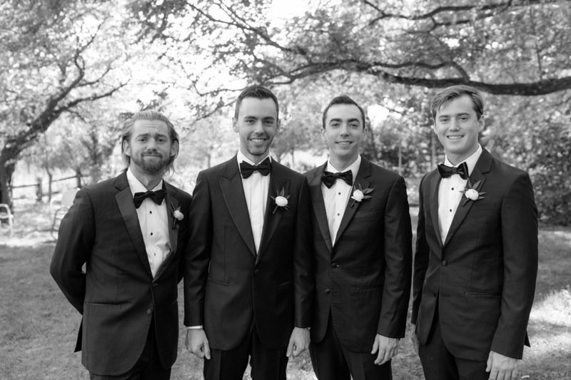 Four men in tuxedos standing outdoors in front of trees, smiling for a photo.