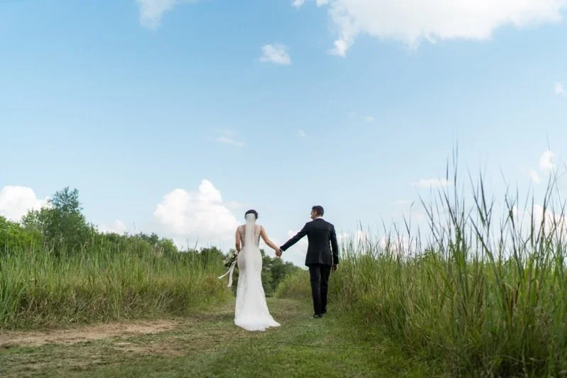 A bride and groom holding hands and walking down a grassy path in a natural outdoor setting.