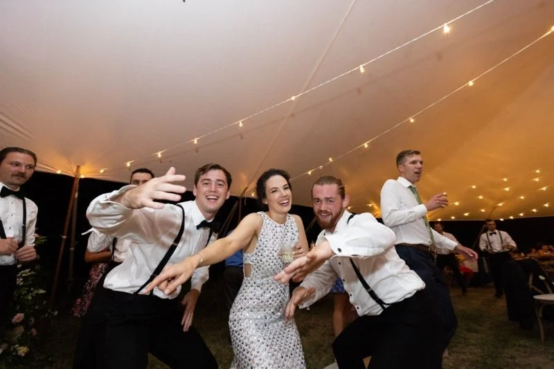 Group of people dancing and celebrating at a fancy event under string lights, with some holding drinks and smiling.