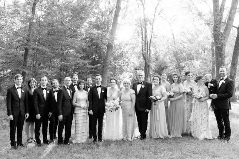 A large wedding party with men in tuxedos and women in various dresses standing outdoors in a wooded area.
