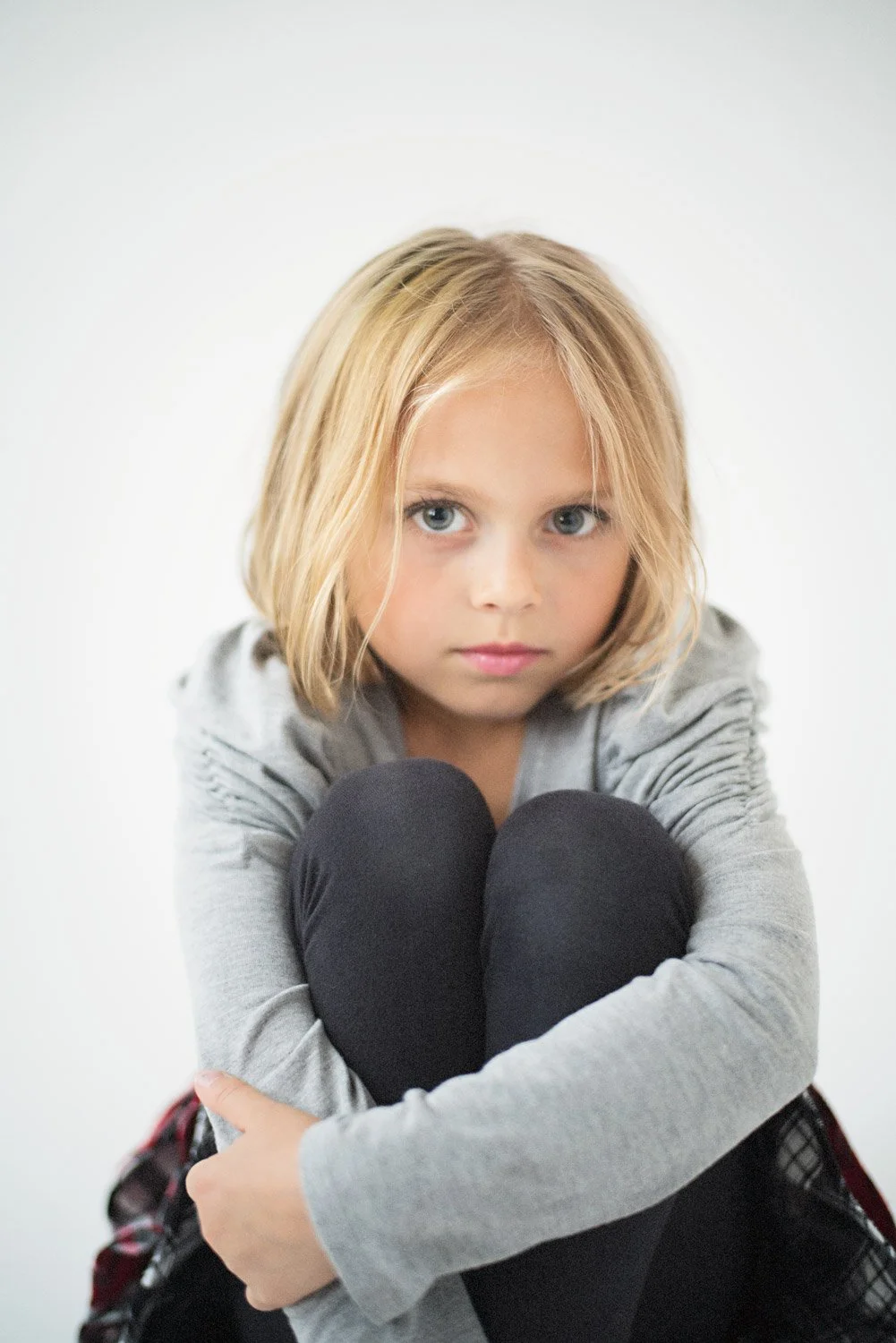 A young girl with blonde hair and blue eyes sits with her legs pulled to her chest, looking directly at the camera against a plain white background.
