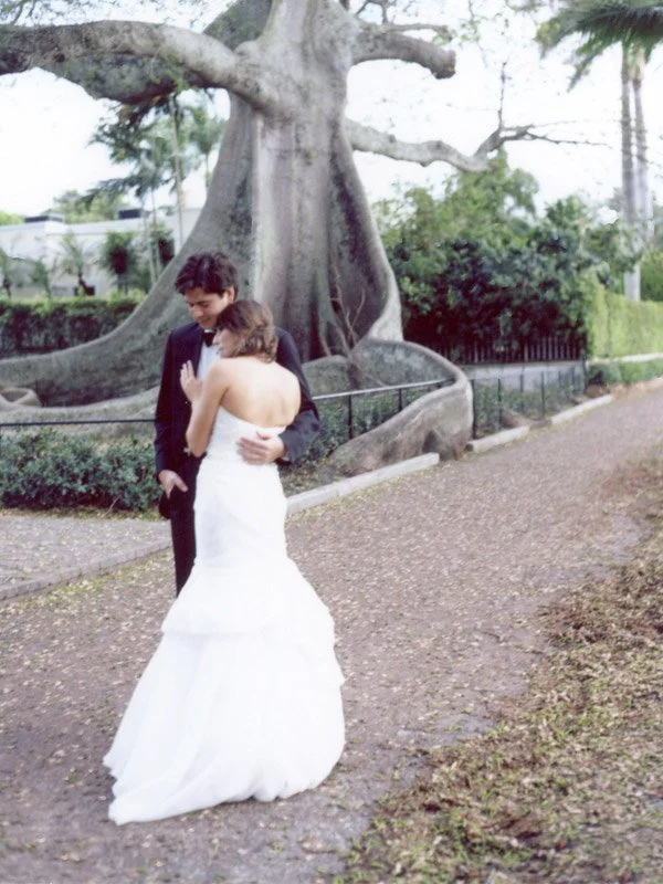 A bride and groom standing together outdoors near a large tree, sharing a tender moment.