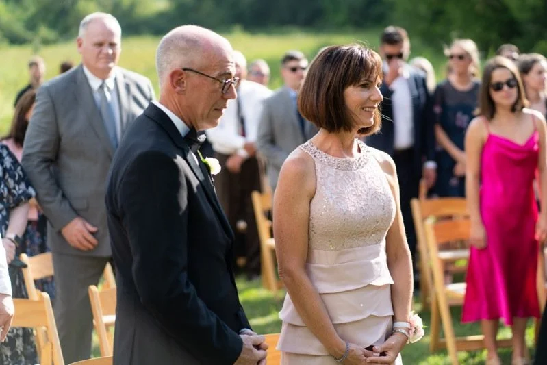 A woman with a bob haircut wearing a light-colored dress is smiling during an outdoor wedding ceremony. An older man in a tuxedo stands beside her, with guests seated and standing in the background.