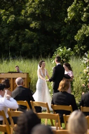 A couple getting married outdoors on a sunny day, holding hands and smiling at each other, surrounded by seated guests.