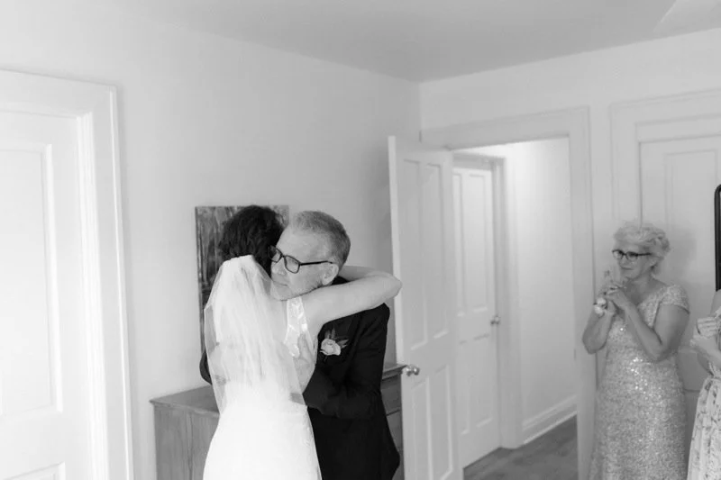 A bride hugging an older man, possibly her father, in a room with two women watching in the background.