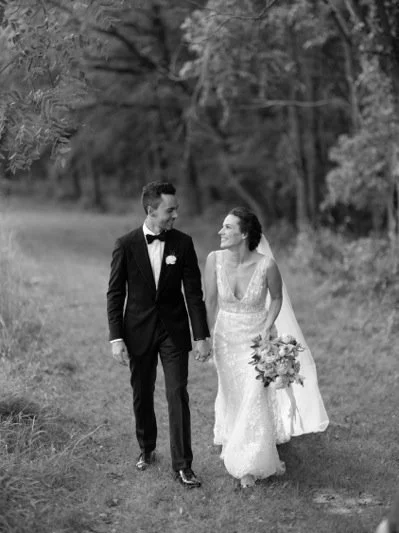Black and white photo of a bride and groom walking hand in hand outdoors, with trees and grass in the background.