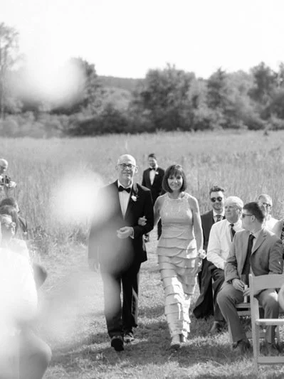 A wedding ceremony outdoors in a grassy field, with guests seated and a couple walking down the aisle. The groom is in a tuxedo, and the bride wears a ruffled dress.