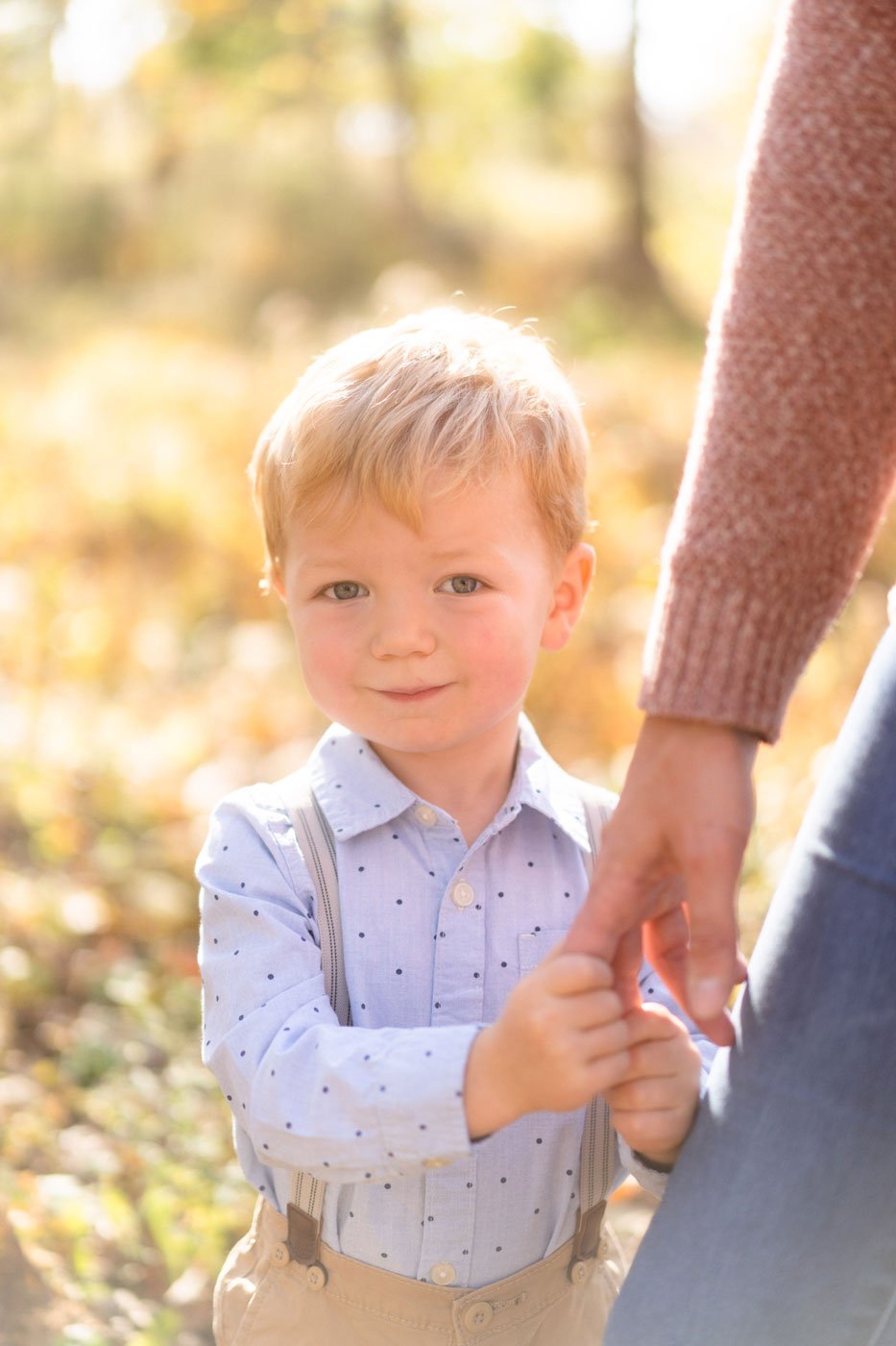 A young boy with blonde hair and blue eyes holding hands with an adult outside during daytime with a blurred autumn background.