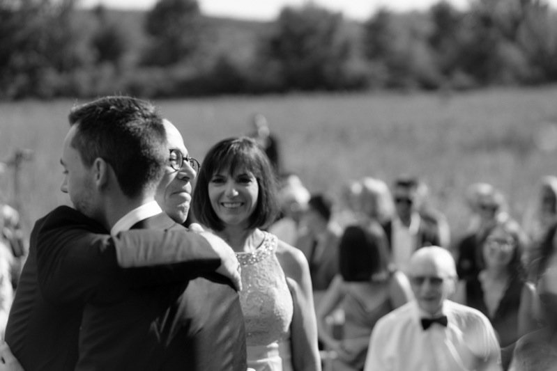 A black-and-white photo of a couple dancing outdoors at a wedding, with a woman smiling and a man with glasses and a suit, and guests in the background.
