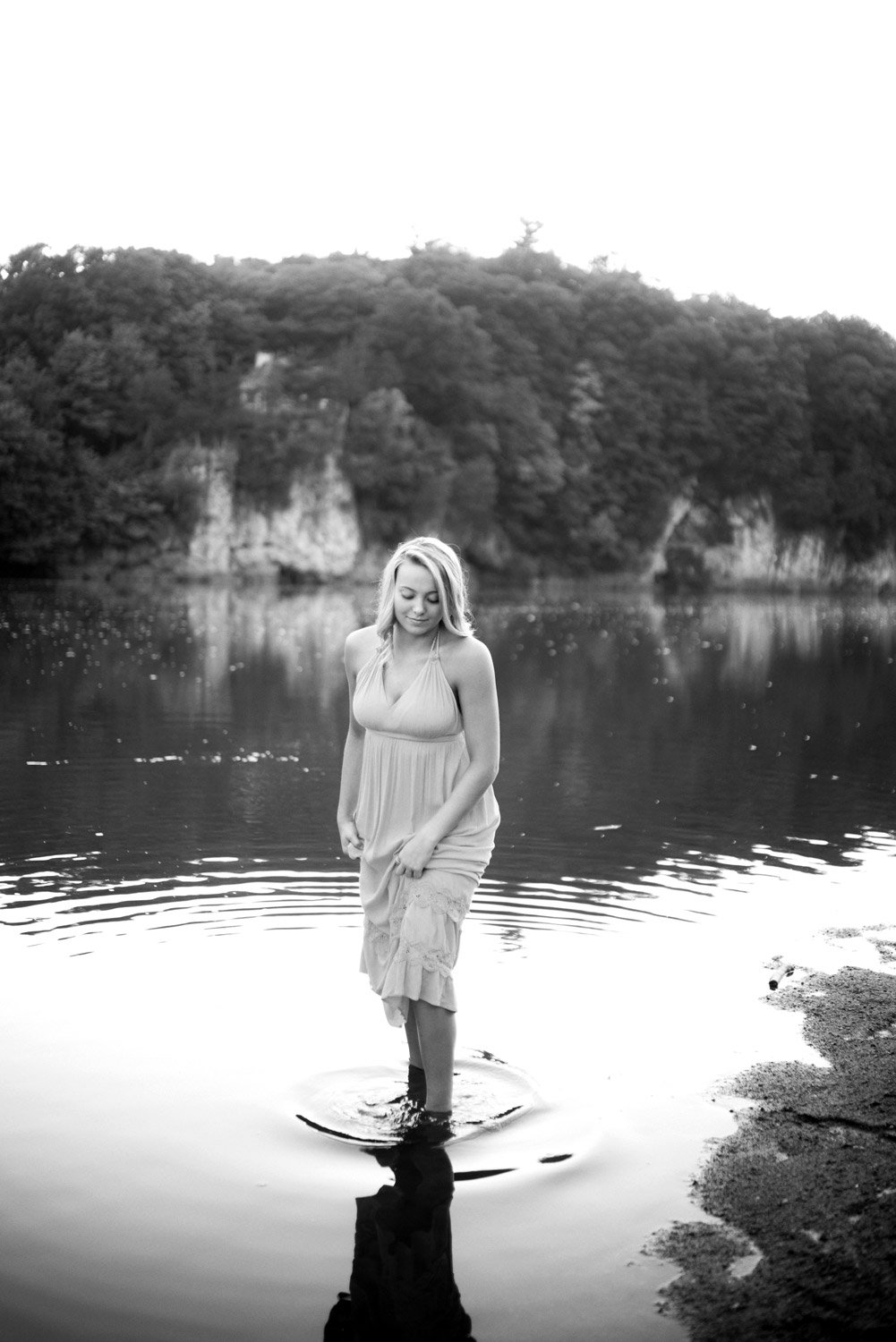 A woman in a flowing dress stands in water near a shoreline, with trees and cliffs in the background, captured in black and white.