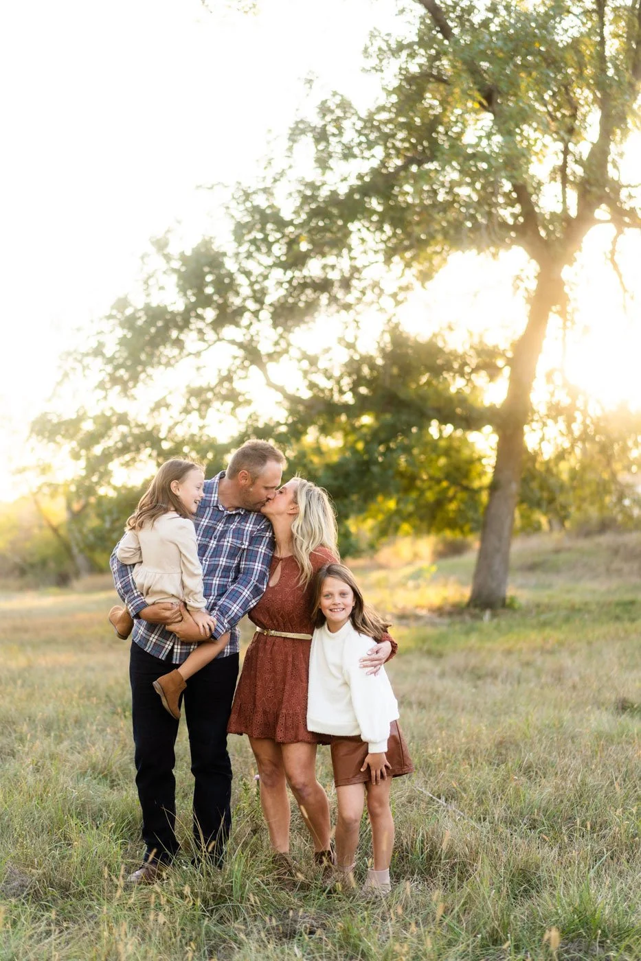 A family of four standing outdoors in a grassy field during sunset, with trees in the background. The father and mother are kissing, the father holding a young girl, and the older girl standing beside the mother, smiling.