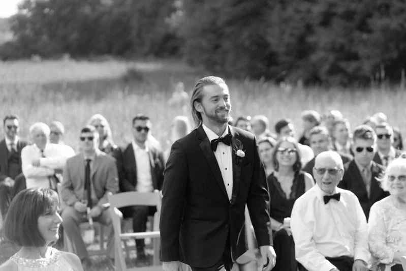 A black-and-white photograph of a wedding ceremony outdoors with a groom in a suit and bow tie smiling, surrounded by seated guests in formal attire.