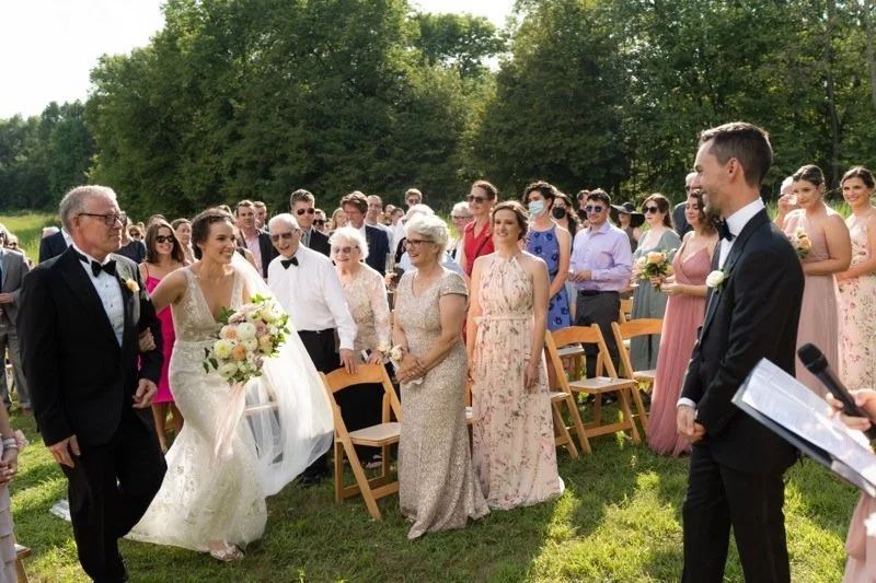 Bride walking down aisle with her father at an outdoor wedding, others seated and standing on green grass, trees in background, sunny day.