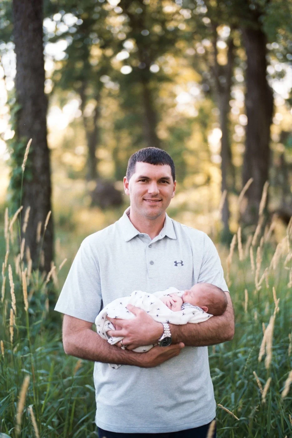 A man holding a newborn baby wrapped in a blanket outdoors in a field with tall grass and trees in the background during golden hour.