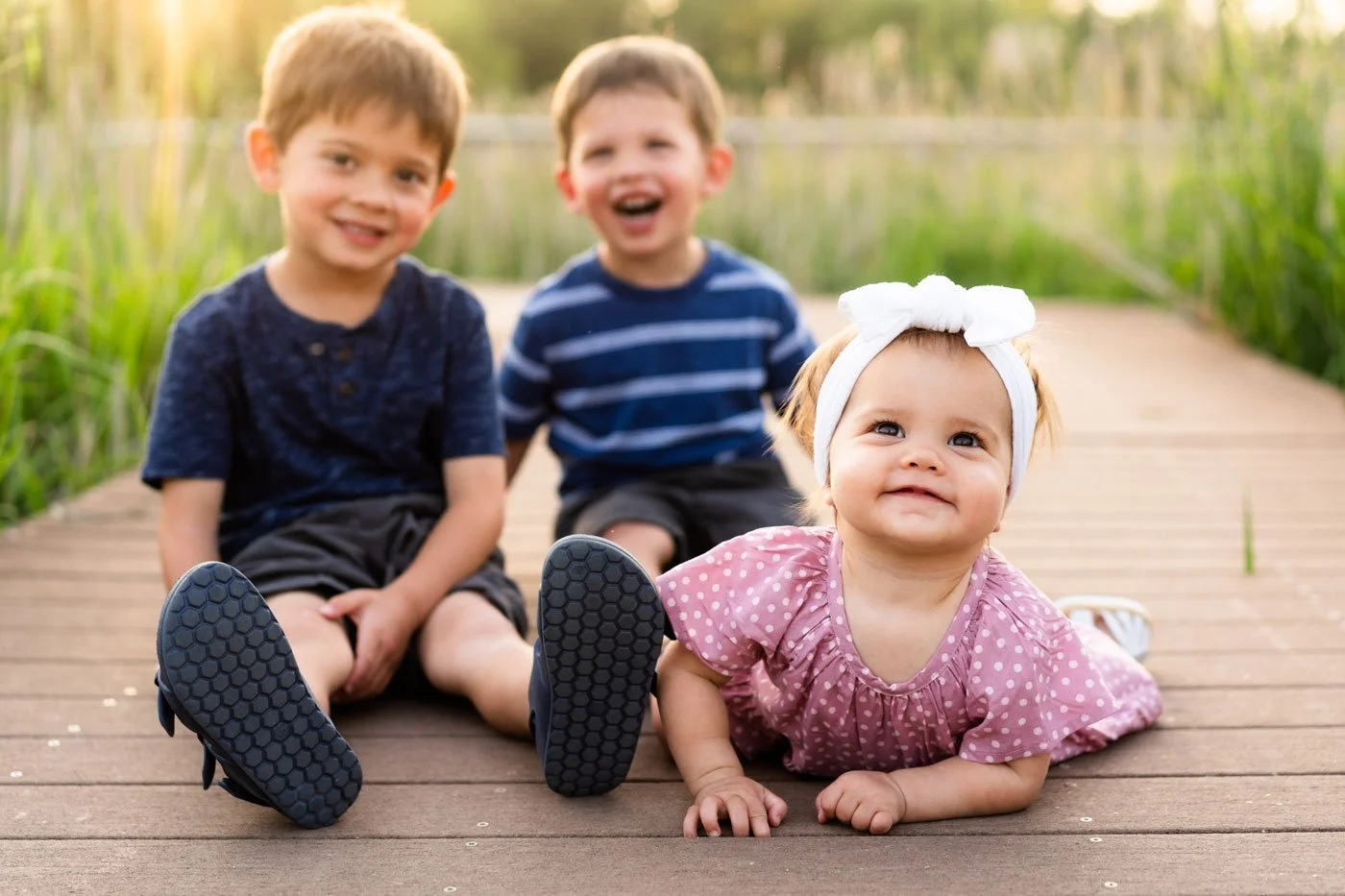 Three young children sitting on a wooden dock near a pond, smiling and playing outdoors on a sunny day.