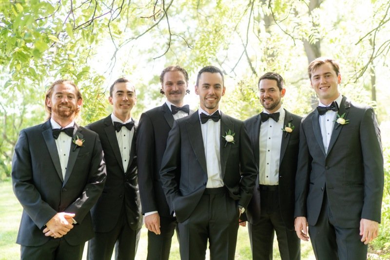 Six men dressed in black tuxedos with bow ties, standing outdoors under trees with green leaves. They are smiling, facing the camera, and appear to be at a formal event or wedding.