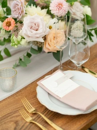 Elegant table setting with a floral centerpiece, wine glasses, gold flatware, a white plate, and a pink napkin, with a white tablecloth.