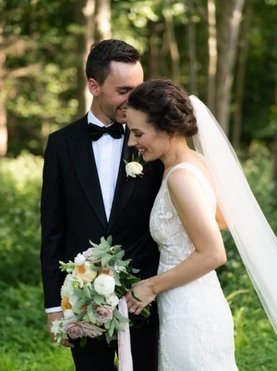 A bride and groom standing close together outdoors, smiling, with the bride holding a bouquet of flowers, surrounded by green trees.