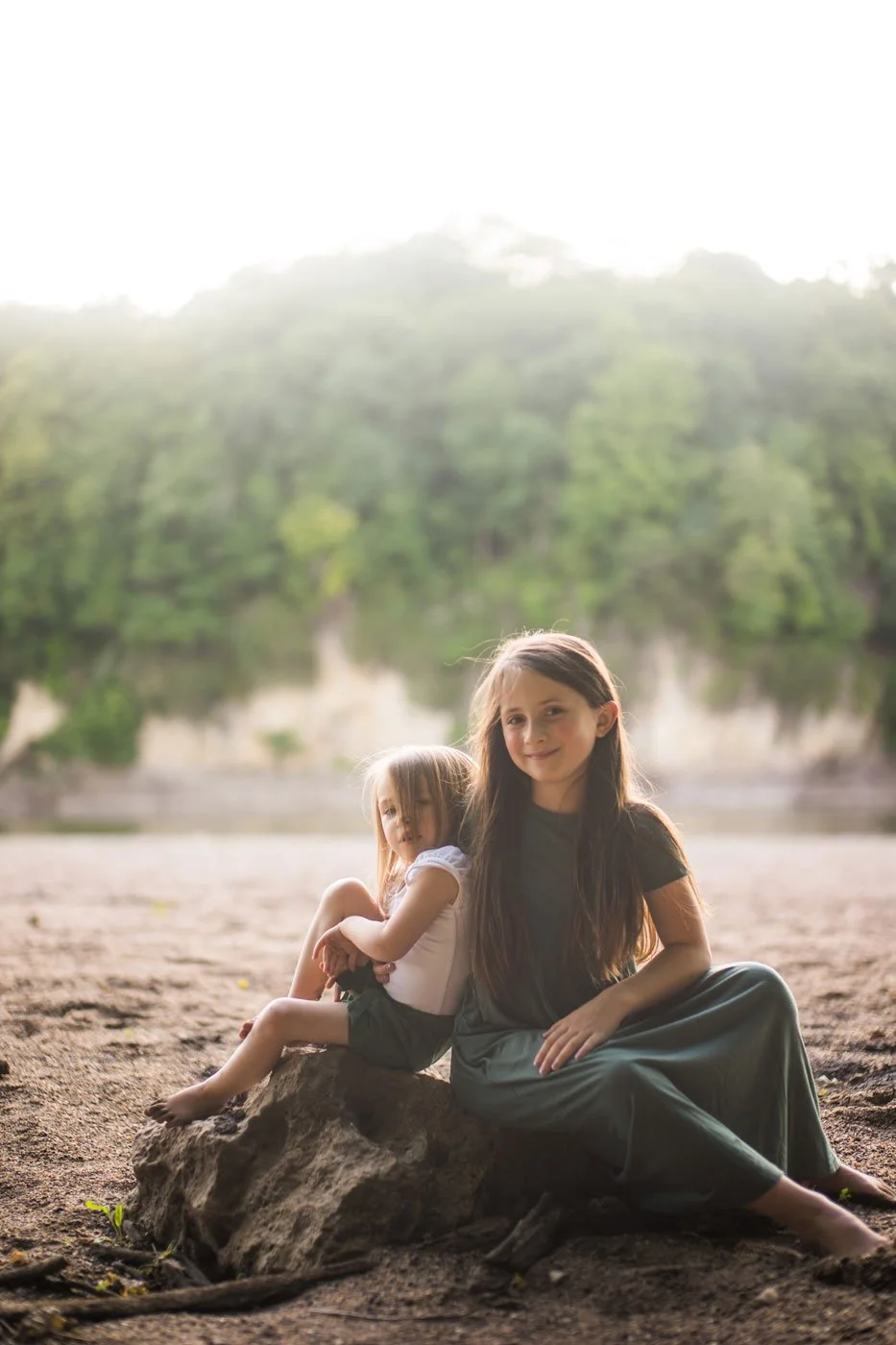Two young girls sitting on a rock outdoors with a blurred green forest in the background, lit by soft natural light.