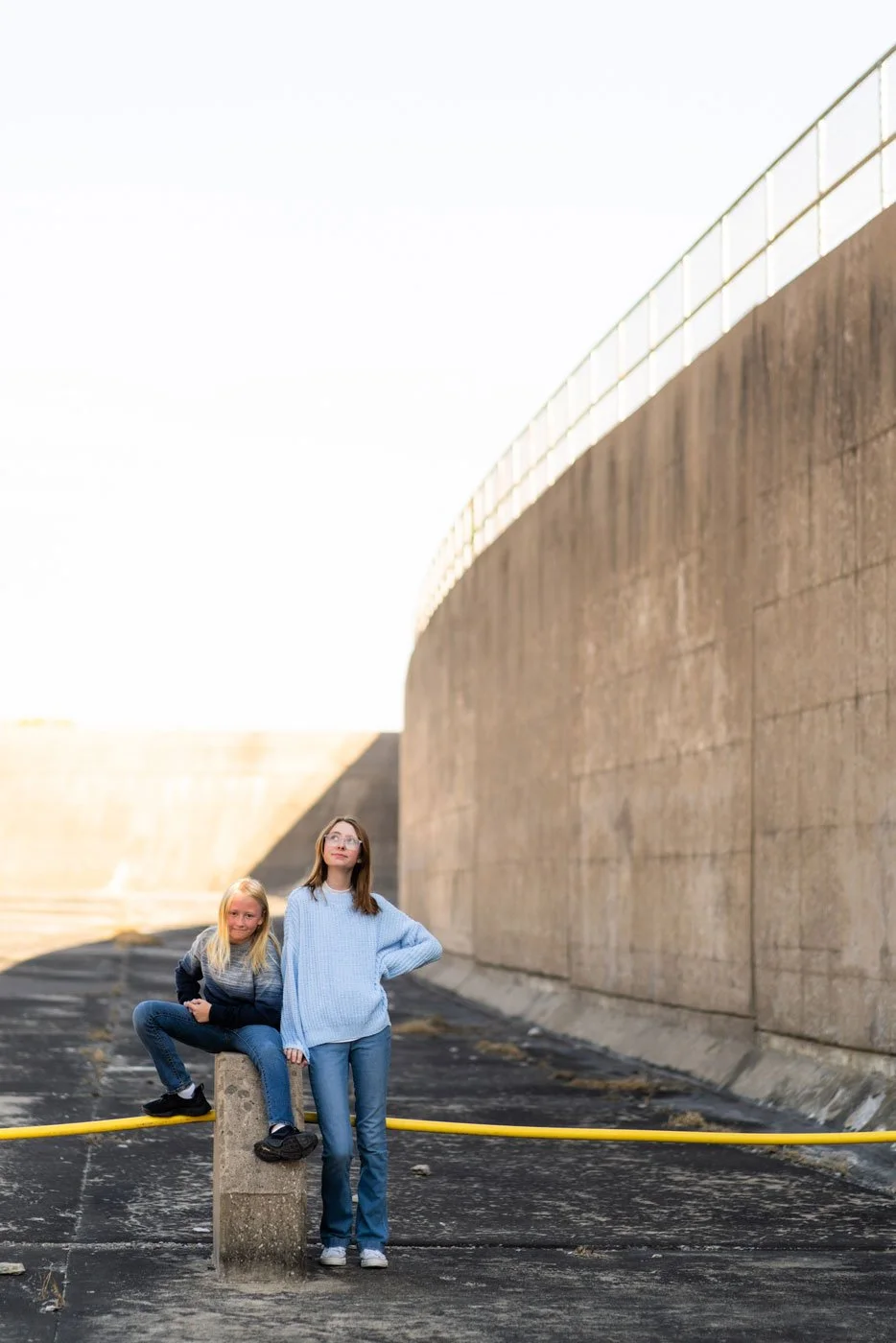 Two girls pose near a large concrete structure with a curved wall, one sitting on a concrete block and the other standing, both looking into the distance.