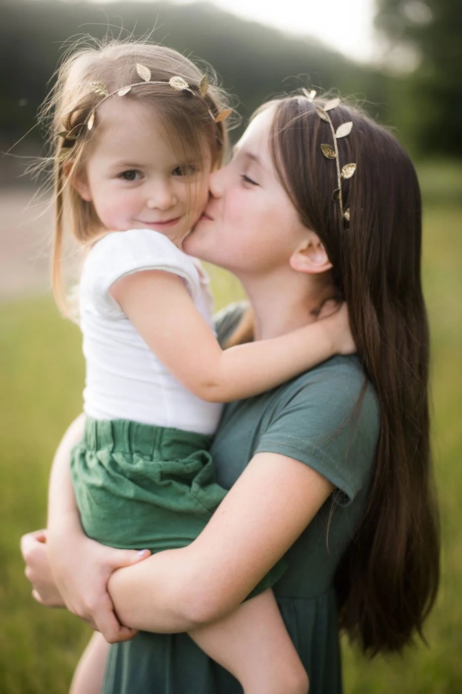 A woman is holding a young girl, who is kissing her cheek. Both are wearing leaf-themed headbands, and are outside in a grassy area with trees in the background.