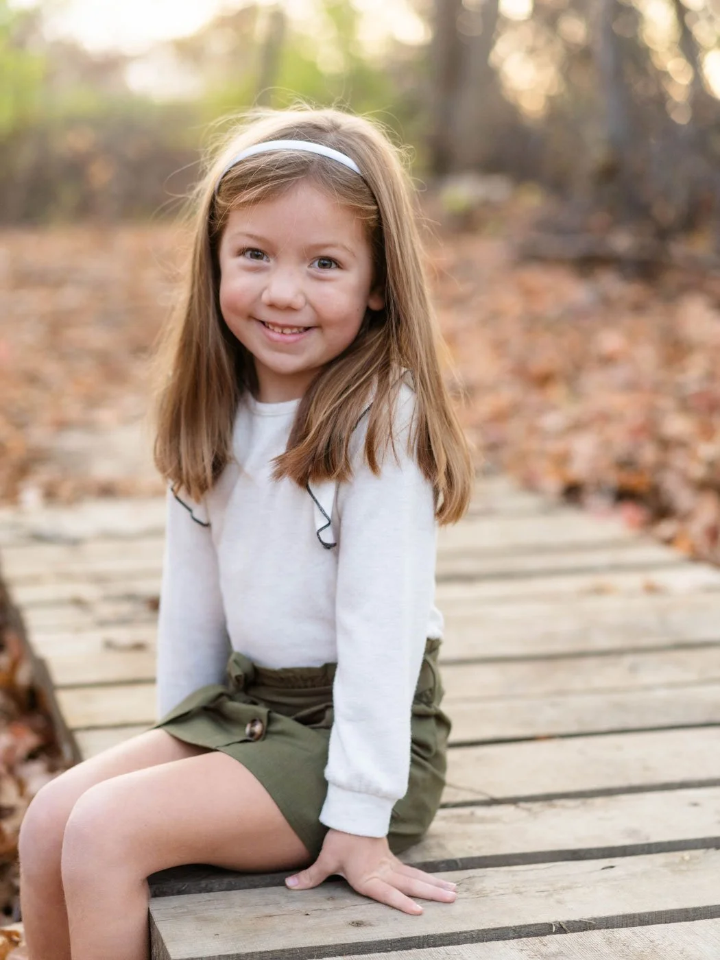 A young girl with long light brown hair, wearing a white long-sleeve shirt, olive green shorts, and a white headband, sitting on a wooden bridge in an outdoor wooded area during autumn.