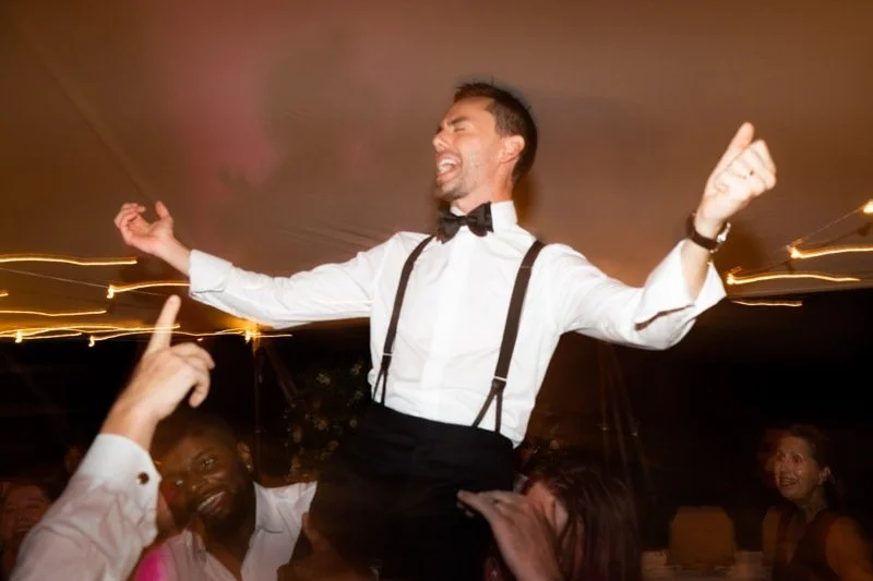 A man in a tuxedo with suspenders and a bow tie, singing or celebrating enthusiastically while being lifted or surrounded by others at a party or celebration.
