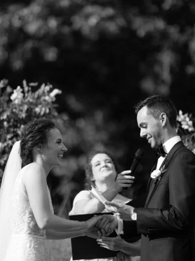 Bride and groom during their wedding ceremony, with a woman holding a microphone and a paper, outdoors.