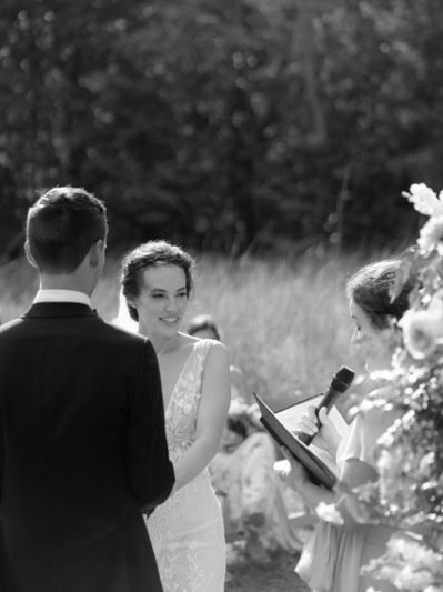 A wedding ceremony outdoors with a bride and groom facing each other, someone officiating, and flowers, in black and white.