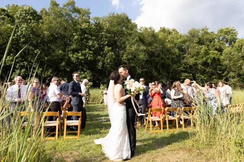Bride and groom kissing during outdoor wedding in a grassy field, with wedding guests standing and seated nearby, surrounded by trees and tall grass.