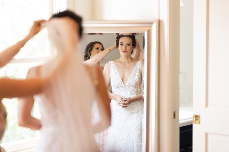 A woman in a wedding dress looking at herself in a mirror while another woman helps her with her hair.