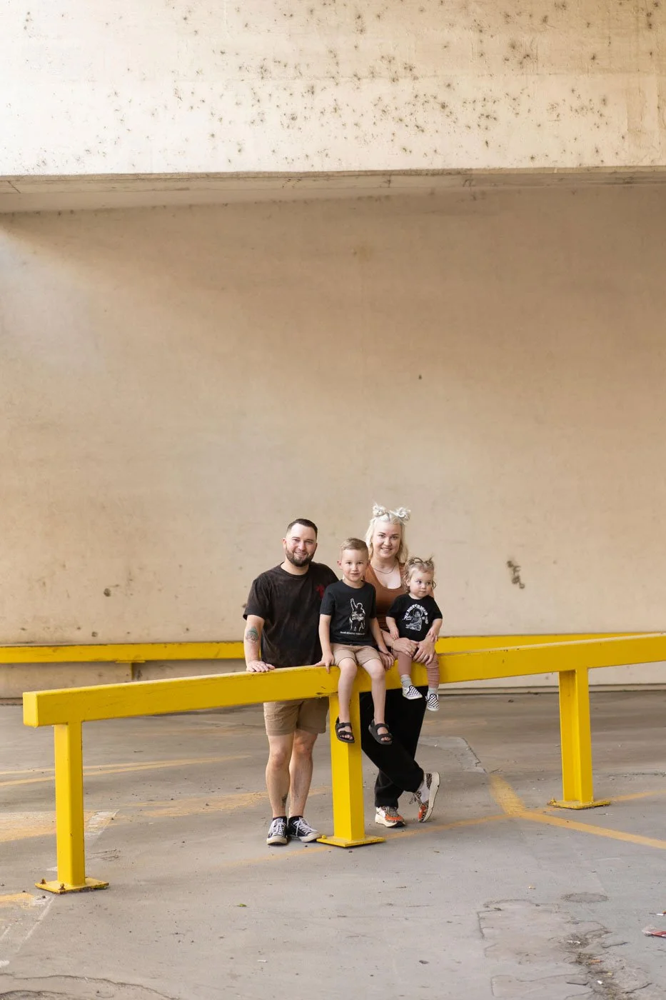 Family of four sitting on yellow parking lot barrier in an indoor parking garage, smiling at the camera.