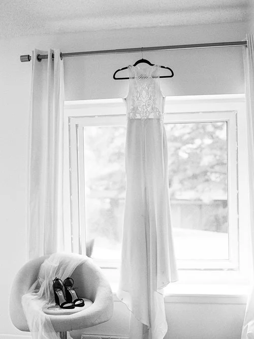 Wedding dress hanging on a curtain rod in front of a window with a veil and a pair of black high-heeled shoes on an armchair