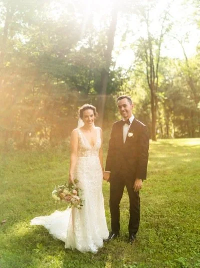 A bride and groom standing outdoors on grass with trees in the background, holding hands, at sunset.