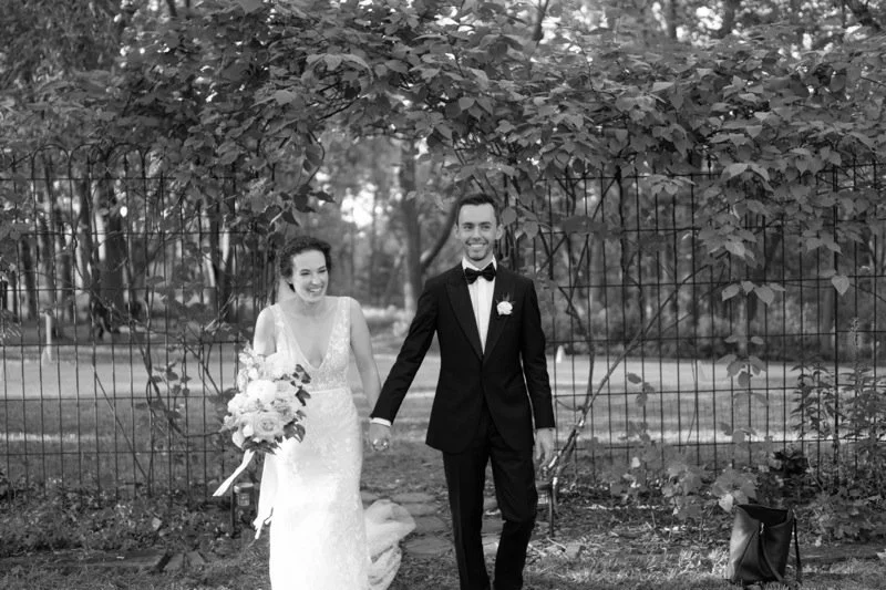 A black and white photo of a bride and groom walking outdoors, holding hands. The bride is wearing a lace wedding dress and holding a bouquet, while the groom is dressed in a tuxedo with a bow tie. They are smiling and appear happy.