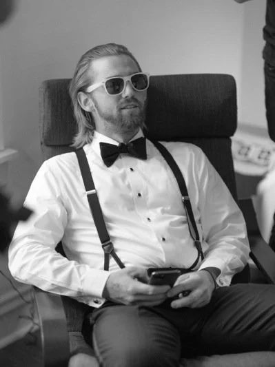 Man with long hair and beard wearing sunglasses, tuxedo with bow tie and suspenders, sitting on an office chair, holding a smartphone.