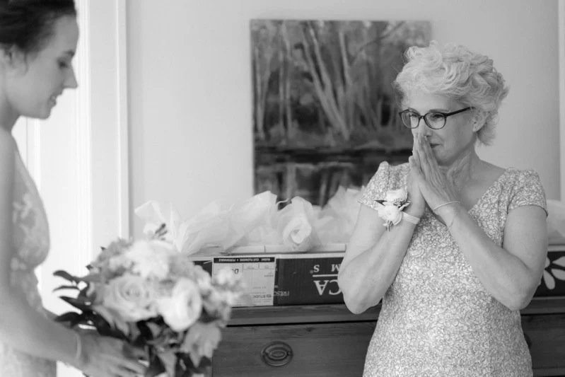 An elderly woman with white hair and glasses, dressed in a sparkly dress, stands with her hands clasped in prayer in front of a young woman holding a bouquet of flowers, in a room with a painting and some gift boxes.