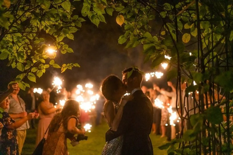 Couple dancing closely at night during a celebration with sparklers held by guests in the background, surrounded by trees.