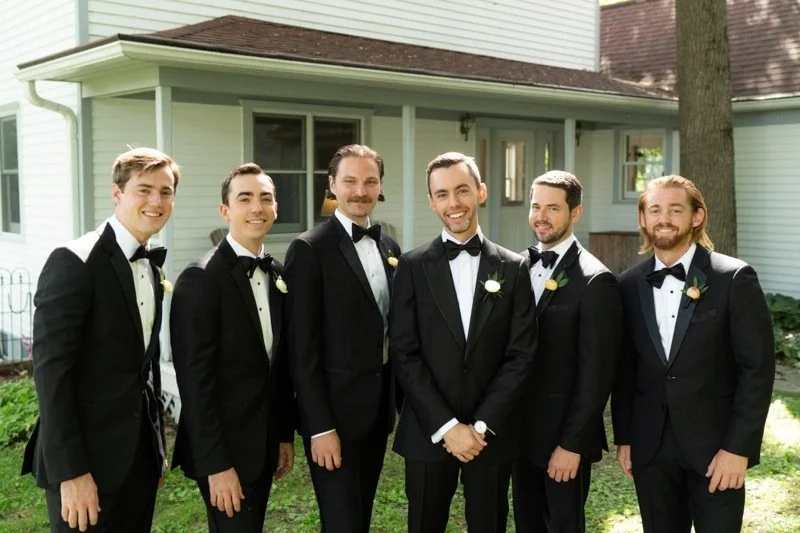 Six men in tuxedos with bow ties and boutonnières standing outdoors in front of a house, smiling.
