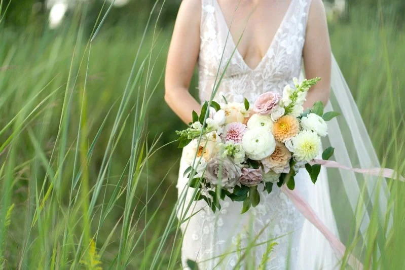 A bridesmaid in a white lace wedding dress holding a large bouquet of pastel-colored flowers, standing among tall green grass.