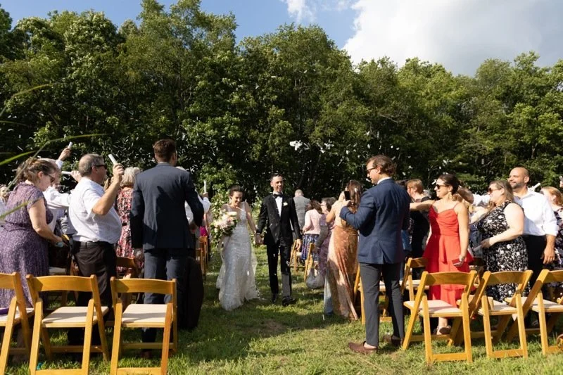 Wedding ceremony outdoors with bride and groom walking between rows of seated guests, surrounded by greenery and clear blue sky.