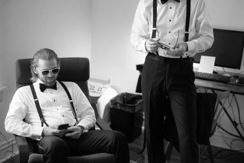 Two men in formal attire, one sitting with sunglasses and a beard, the other standing with a notepad in a room with a computer and a trash bin.