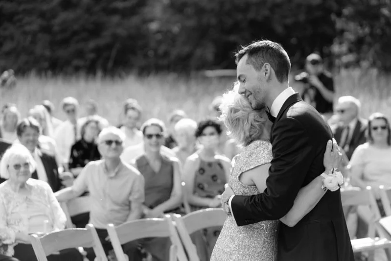 A groom hugging an older woman during a wedding ceremony outdoors, with guests seated and standing in the background.