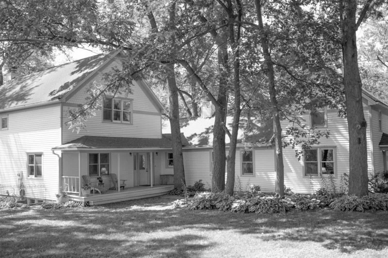 A two-story house with a porch, surrounded by large trees and a grassy yard.