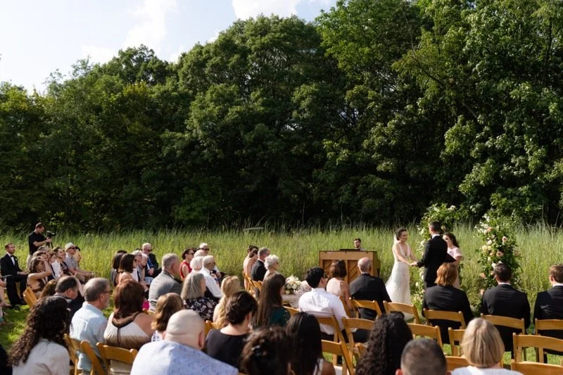 Outdoor wedding ceremony with bride and groom exchanging vows, surrounded by seated guests, with lush green trees in the background.