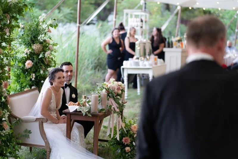 A wedding reception outdoors with a bride and groom sitting on a vintage bench decorated with flowers, while a man in a tuxedo stands in the foreground. In the background, there are guests and a table with drinks.